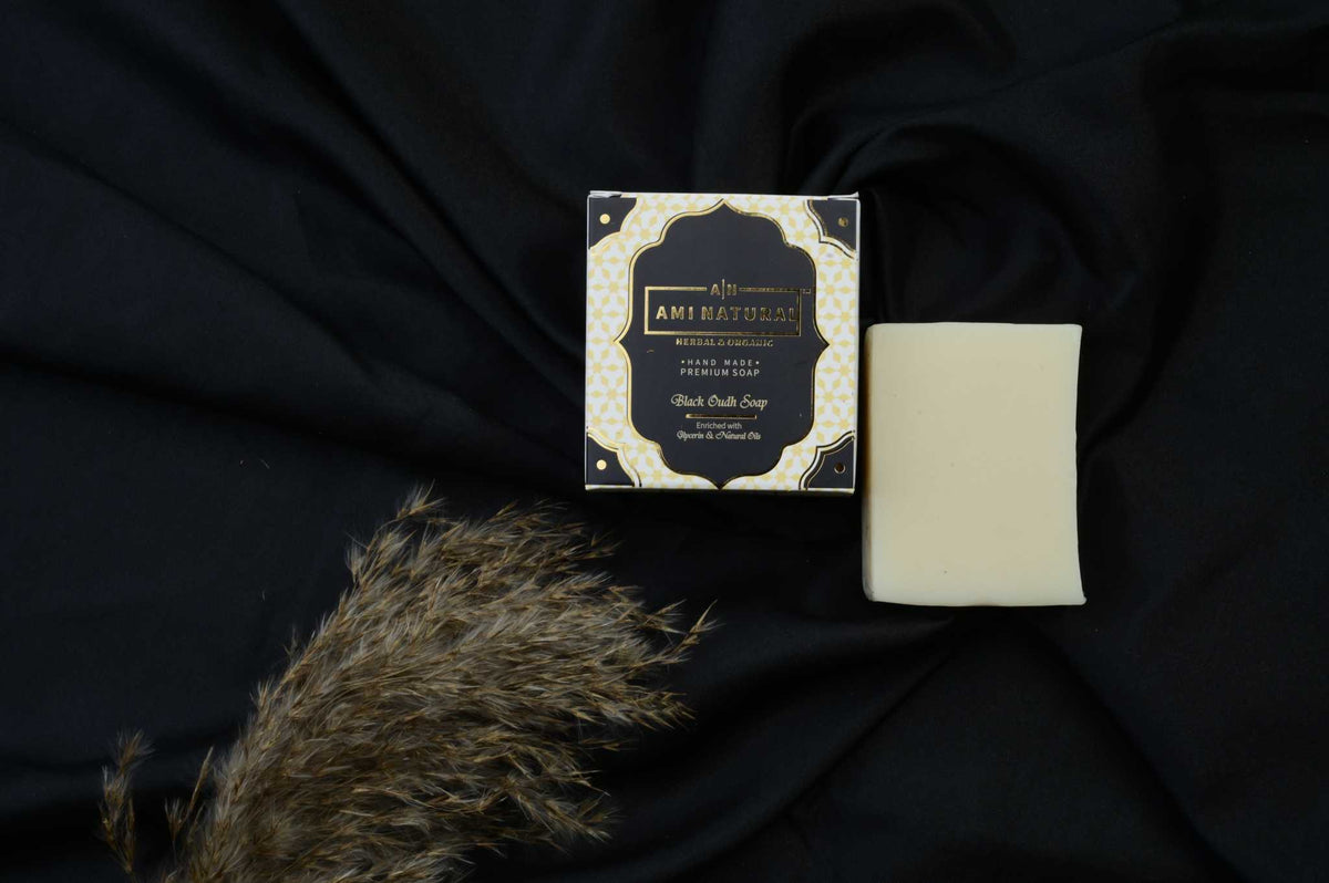 AMI NATURAL BLACK OUDH SOAP with elegant black and gold packaging alongside a plain off-white soap bar on black fabric