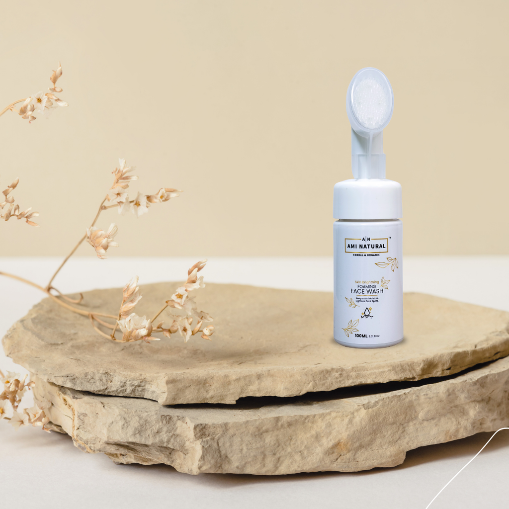 Ami Natural Brightening foaming face wash in white bottle with foam brush cap on natural stone slabs with dried flowers